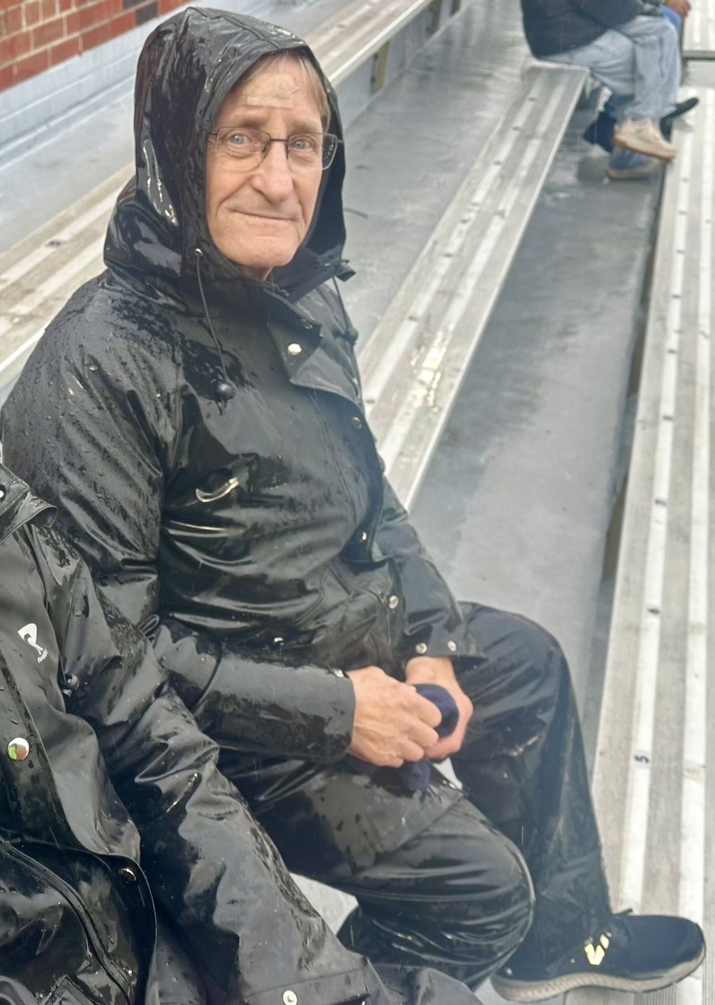 A man sitting on a bleacher with a black rain jacket, black rain pants, and black sneakers.